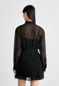 Črna nabrana obleka z prosojnimi dolgimi rokavi in pasom, ki vključuje zadnji yoke in subtilne gumbke na manšeta.