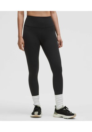 Donna che indossa leggings neri a vita alta, calzini bianchi arricciati e scarpe sportive nere, in piedi davanti a uno sfondo semplice.