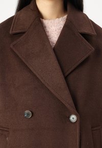 Hnědý vlněný kabát s klopami, dvouřadým zapínáním a šedými knoflíky. Obsahuje měkkou, texturovanou vnitřní vrstvu, která je viditelná.
