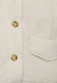 Miękka, kremowa tkanina sherpa z dwoma bursztynowymi guzikami i detalem kieszeni z klapką na odzieży.