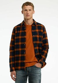 Geruit flanellen shirt in marineblauw en oranje met een knoopsluiting aan de voorkant, gedragen over een oranje T-shirt met ronde hals. Blauwe jeans maken de look compleet.