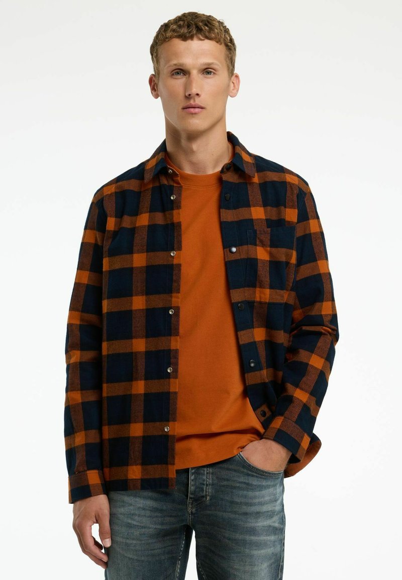 Geruit flanellen shirt in marineblauw en oranje met een knoopsluiting aan de voorkant, gedragen over een oranje T-shirt met ronde hals. Blauwe jeans maken de look compleet.