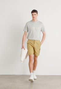 Tricou gri deschis cu text "Barbour International", pantaloni scurți bej și adidași albi. Modelul ține un pulover de culoare deschisă. Fundal neutru.