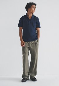 Temno modra polo majica s kratkim ovratnikom, kombinirana z ohlapnimi, svetlo olivno zelenimi hlačami in temno rjavimi čevlji. Preprost dizajn, teksturirana tkanina.