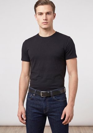 Jeune homme portant un t-shirt noir uni à manches courtes, un jean bleu foncé et une ceinture noire, debout devant un fond clair uni.