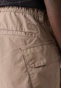 Tan cargo shorts made of lightweight fabric with a textured pattern. Features stitched accents and a small logo label on the back pocket.