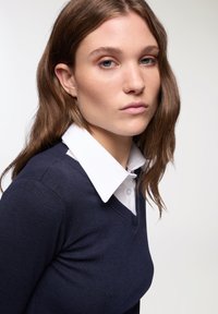 Pullover blu navy con scollo a V indossato sopra una camicia bianca con colletto. Presenta una texture liscia e una silhouette aderente. I capelli cadono in morbide onde.