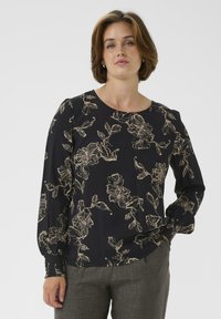 Donna vestita con una blusa nera con motivi floreali beige e maniche lunghe, abbinata a pantaloni grigi, in piedi di fronte a uno sfondo neutro.