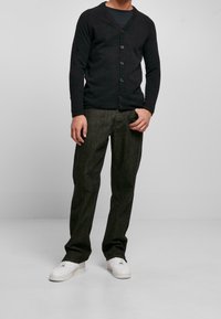 Svart stickad cardigan med v-ringning och knappstängning, tillsammans med mörkgrå byxor och vita sneakers, stående mot en neutral bakgrund.