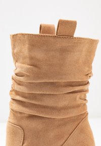 Bottine décontractée en daim beige avec une texture froncée, dotée de deux languettes en tissu au niveau du col et d'un bout arrondi lisse.