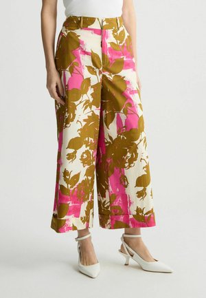Kobieta w spodniach z szerokimi nogawkami w brązowe kwiatowe i różowe abstrakcyjne wzory, połączonych z białymi butami na obcasie i białym topem.