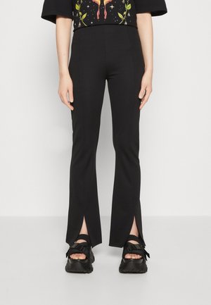 Pantalones negros ajustados con aberturas frontales en el bajo, combinados con sandalias negras y una camiseta gráfica parcialmente visible sobre una persona de pie.