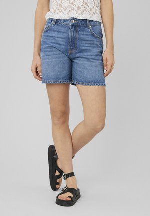 Personne portant un short en jean bleu, un haut sans manches en dentelle blanche, et des sandales à plateforme noires, debout jambes croisées devant un fond gris clair.