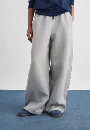 Persona con pantalones deportivos grises holgados con el logo de Adidas, una chaqueta azul marino y zapatillas azules de pie sobre una alfombra azul.