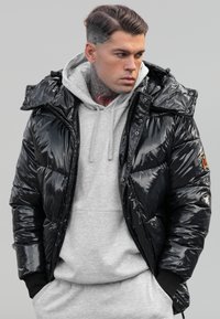 Doudoune noire brillante avec une capuche, fermeture éclair et poignets élastiques, portée par-dessus un sweat à capuche gris clair, sur un fond neutre.