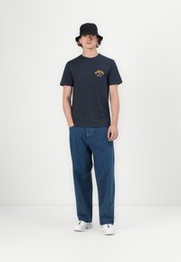 Navy cotton t-shirt with yellow "Florida" graphic on the chest, paired with loose blue denim jeans and white sneakers. Bucket hat worn.