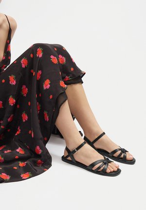 Schwarze Leder-Sandalen mit Kreuzriemen, flachem Absatz und verstellbarem Knöchelriemen. Kombiniert mit einem schwarzen Blumenkleid, das mit roten und rosa Blumen verziert ist.