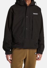 Personne portant une veste à capuche Timberland noire avec boutons-pression et un pantalon Timberland marron.
