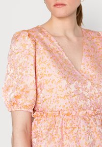 Vestido de mangas bufantes em rosa claro com padrões florais dourados, apresentando um design de decote em V e cintura franzida, feito de um tecido texturizado e sedoso.