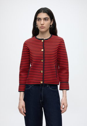 Donna con i capelli lunghi e scuri che indossa una giacca abbottonata a righe orizzontali rosse e nere e jeans blu scuro, in piedi davanti a uno sfondo semplice.