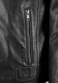 Svart läderjacka med en framträdande dragkedja med en silvrig dragflik, strukturerad yta och en ytterligare rektangulär lapp nära dragkedjan.