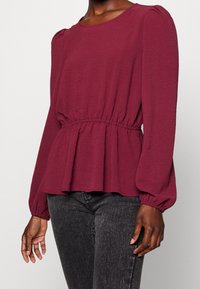 Kvinna i en långärmad vinröd blus med samlad midja och puffade axlar, i kombination med mörkgrå jeans.