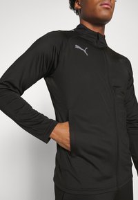 Musta vetoketjullinen takki, joka on valmistettu sileästä, venyvästä kankaasta. Takissa on korkea kaulus, sivutaskut ja pieni harmaa Puma-logo rintamuksessa.