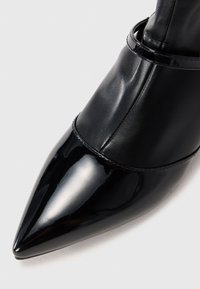 Scarpa nera con punta appuntita, caratterizzata da una tomaia in pelle verniciata lucida, con un cinturino alla caviglia in pelle opaca e dettagli di cucitura sottili.