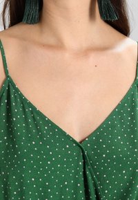 Green, polka-dotted garment with a v-neck design, featuring thin straps and a soft texture. Large, textured green tassel earrings.