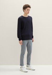 Námornícky pruhovaný pletený sveter s dlhými rukávmi, v kombinácii so svetlošedými slim-fit nohavicami a bielymi teniskami, na jednoduchom pozadí.