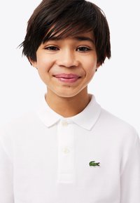 Lächelnder kleiner Junge mit kurzen dunklen Haaren, der ein weißes Poloshirt mit einem grünen Krokodil-Logo auf der Brust trägt.