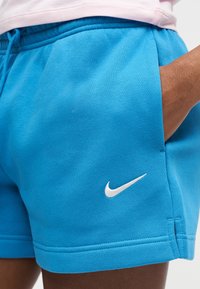Άτομο που φοράει φωτεινά μπλε σορτς Nike με λευκό λογότυπο, το χέρι τοποθετημένο χαλαρά στην τσέπη, ορατό κοντά στη μέση και την περιοχή του μηρού.
