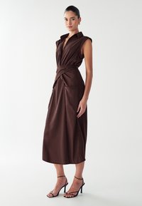 Robe marron sans manches avec un col, taille ceinturée, deux poches latérales et une silhouette fluide. Modèle associé à des sandales noires.