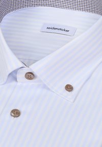Gros plan d'une chemise blanche à rayures bleu clair, boutons beiges, avec une étiquette de marque "seidensticker" visible à l'intérieur du col.