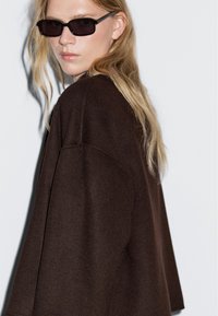 Donna con lunghi capelli biondi che indossa occhiali da sole scuri e un cappotto largo di colore marrone scuro, appoggiata su uno sfondo chiaro, che guarda di lato.