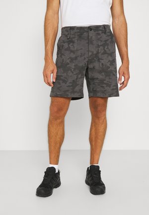 Homme portant un short camouflage gris foncé, des chaussettes blanches, des baskets noires et une chemise blanche sur un fond blanc uni.