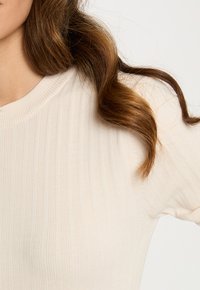 Primer plano del hombro y cuello de una persona que lleva un suéter de canalé color crema y tiene el cabello castaño suelto y ondulado.