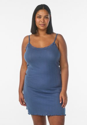 Kvinde med skulderlangt sort hår iført en blå ribstrikket ærmeløs kjole med blonder, stående foran en ensfarvet hvid baggrund.