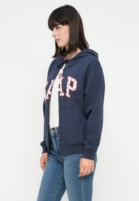 Žena u tamnoplavoj GAP hoodie s cvjetnim ispisanim slovima, bijelom majicom i plavim trapericama, stoji u profilu protiv obične bijele pozadine.