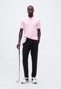 Άνδρας στέκεται κρατώντας μπαστούνι γκολφ, φορώντας ροζ πόλο πουκάμισο, μαύρο παντελόνι με λευκό γάντι γκολφ στην τσέπη και λευκά αθλητικά παπούτσια.