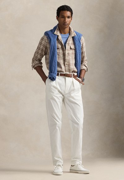 Καρό πουκάμισο σε γήινες αποχρώσεις, φορεμένο κάτω από μπλε πλεκτό πουλόβερ. Λευκά ραμμένα παντελόνια και λευκά αθλητικά παπούτσια ολοκληρώνουν την εμφάνιση.