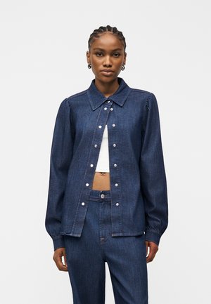 Frau mit geflochtenem Haar, die eine dunkelblaue, geknöpfte Jeansjacke über einem weißen, kurzen Top und passenden Jeanshosen trägt, nach vorne blickend.