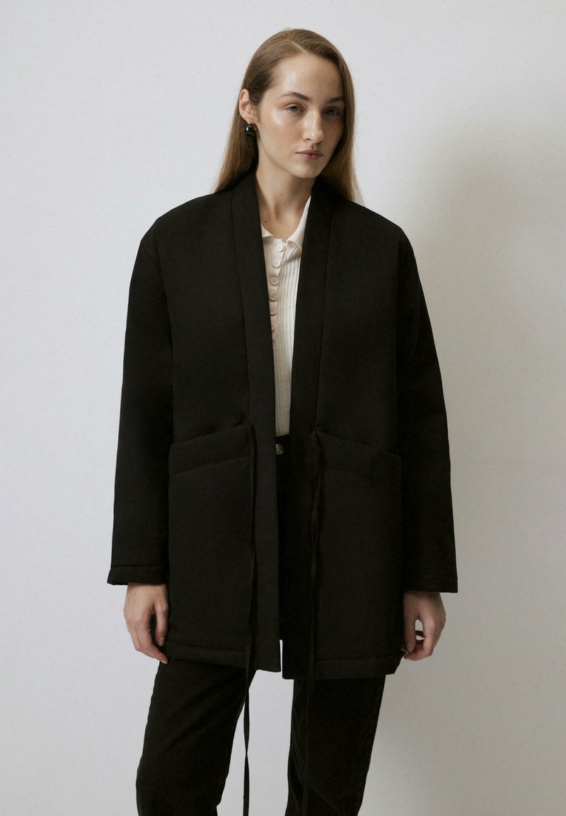 Giacca nera in stile kimono con finitura texture, dotata di chiusura a legaccio, due tasche frontali e maniche larghe. Indossata sopra un top leggero.