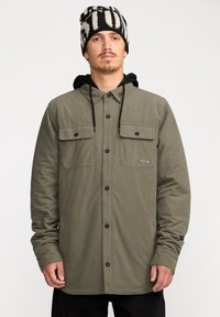 Veste en velours côtelé vert olive avec deux poches poitrine, boutons pression noirs et une capuche. Portée avec un bonnet rayé noir et blanc.