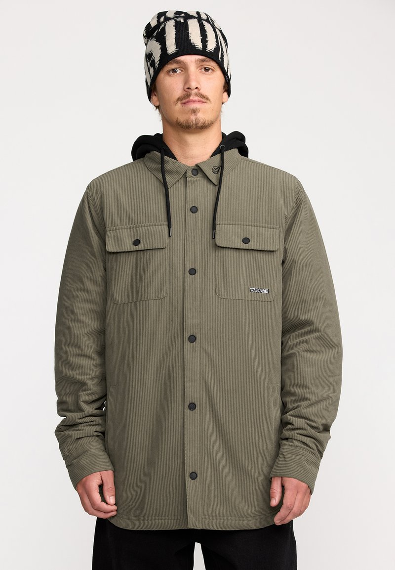 Veste en velours côtelé vert olive avec deux poches poitrine, boutons pression noirs et une capuche. Portée avec un bonnet rayé noir et blanc.