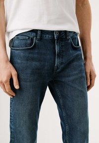 Jeans de mezclilla azul oscuro con un corte recto, cinco bolsillos y una bragueta con botón, combinados con una camiseta blanca, exhibidos en un fondo neutro.