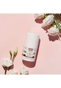 Bottiglia cilindrica bianca di James Read Gradual Tan Sleep Mask Tan per il viso, con dettagli dorati e un'etichetta stampata, circondata da fiori bianchi.