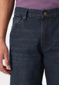 Dark blue denim jeans featuring a slim fit, visible stitching, and a classic metal button closure at the waist. Side pocket detail evident.