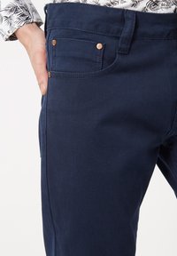 Hombre con pantalones azul oscuro con remaches de cobre, descansando la mano izquierda cerca del bolsillo delantero, vistiendo una camisa blanca con estampado de hojas negras.