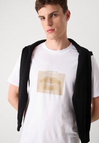 Hvit T-skjorte med et gullmyntegrafikk og "HUGO" tekst, kombinert med en svart genser som henger over skuldrene. Stoffet ser glatt ut.